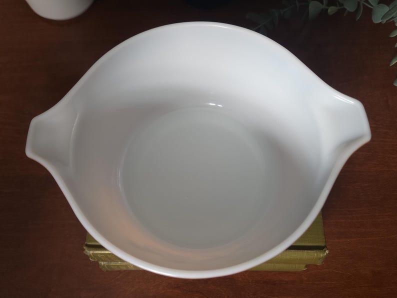 May include: A white, vintage Pyrex mixing bowl with two handles. The bowl has a smooth, glossy finish and a rounded shape. The bowl is sitting on a gold-colored surface.