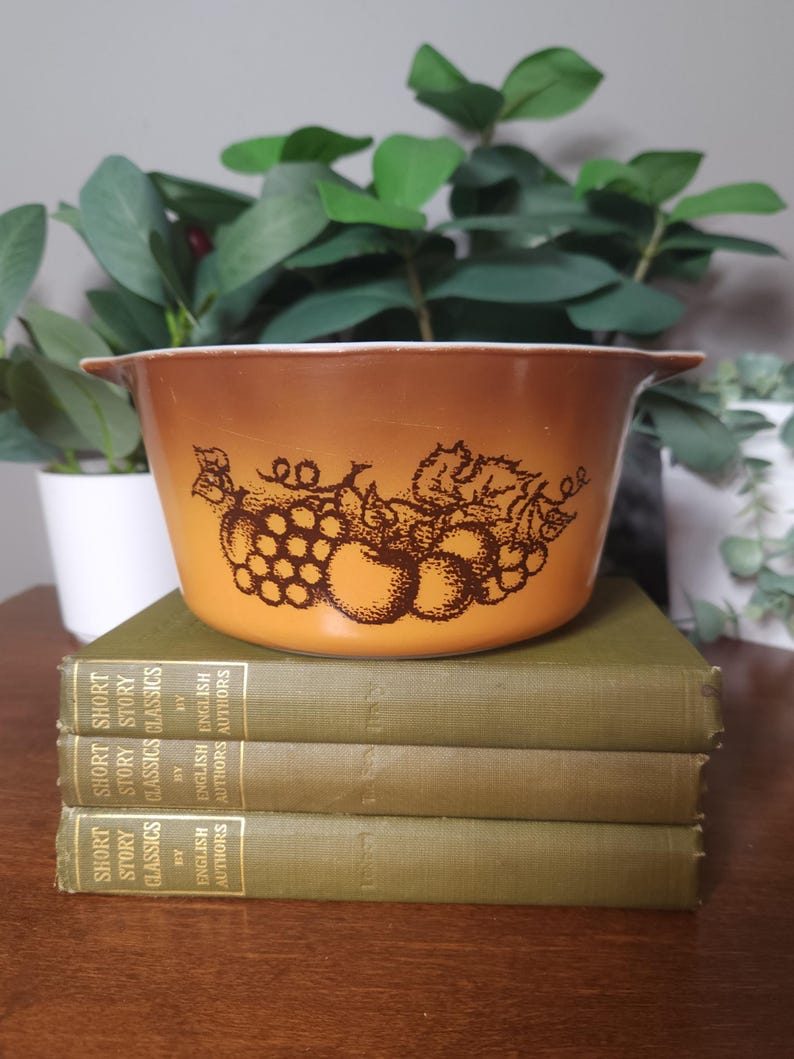 May include: A vintage, round, orange-brown casserole dish with a fruit design. The dish has two handles and is set on a stack of three green books. The books are labeled "Short Story Classics" and "English Authors".