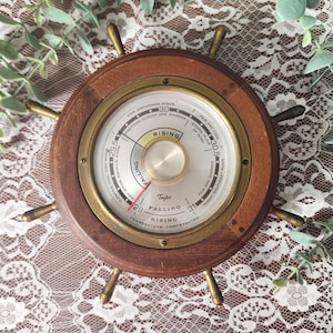 May include: A round wooden barometer shaped like a ship's wheel with a brass rim and center dial. The white face features black text including Taylor, Rising, and Falling, with a red needle pointing toward 29.5, indicating atmospheric pressure.