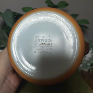 May include: A vintage Pyrex orange and white mixing bowl. The bottom of the bowl is visible, displaying the Pyrex logo and text: "by Corning, Corning N.Y. USA, MAKING AND MICROWAVE."