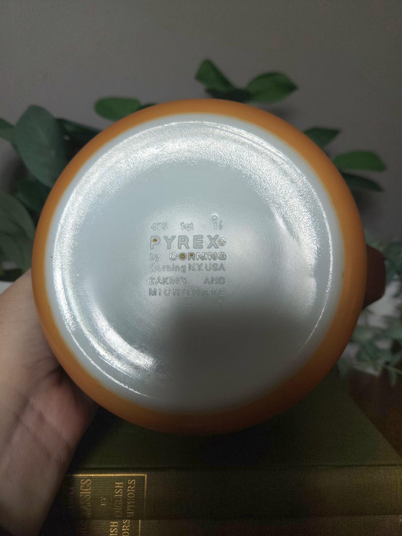 May include: A vintage Pyrex orange and white mixing bowl. The bottom of the bowl is visible, displaying the Pyrex logo and text: "by Corning, Corning N.Y. USA, MAKING AND MICROWAVE."