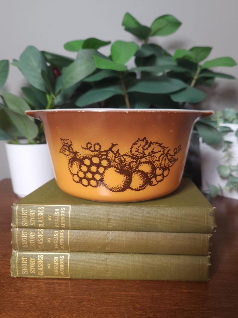 May include: An orange and brown vintage Pyrex bowl with a fruit design, sitting on top of three green books. The bowl has a gradient color and a brown fruit illustration. The books are titled "Short Story Classics".