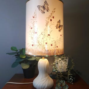 May include: A vintage table lamp with a white ceramic base and a cylindrical lampshade. The lampshade features a cream background with illustrations of butterflies and floral elements. The lamp has a brass-colored trim and a white power cord.