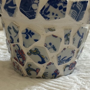 May include: A mosaic-style flower pot with a white exterior and blue and white porcelain shards. The pot's design features floral and geometric patterns, creating a unique decorative item.
