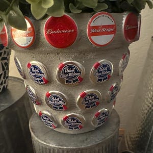 May include: A decorative, gray-colored planter pot adorned with beer bottle caps. The pot is embellished with red and white Budweiser, Red Stripe, and Pabst Blue Ribbon bottle caps. The pot is filled with green leafy plants.