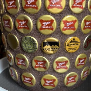 Puede incluir: Una maceta marrón decorada con tapas de botellas de cerveza. Las tapas son principalmente doradas y rojas, con el logotipo "Miller High Life". Otras tapas incluyen "Four Peaks Brewing" y "Bridgeport" brewery.