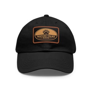 Op de afbeelding: Zwarte baseballcap met een bruine leren patch. De patch heeft een berenpootafdruk en de tekst "NATURE TALKS BACK" in het wit. De cap heeft een gebogen klep en een stoffen bandsluiting.