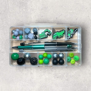 May include: A clear plastic organizer filled with various beads in shades of green, gray, black, and silver. The organizer also contains three green and black pens and three green construction vehicle charms.