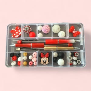Pode incluir: Um organizador de plástico transparente cheio de vários materiais de artesanato. O organizador contém canetas vermelhas, contas decorativas e pingentes, incluindo uma cabeça da Minnie Mouse e formas de borboleta. As contas são em várias cores, incluindo vermelho, rosa, branco, preto e dourado.