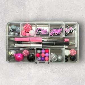 May include: A clear plastic organizer filled with various craft supplies. The organizer contains pink and black pens, colorful beads, and pink construction vehicle charms. The beads are in various colors and textures, including silver, pink, and black.