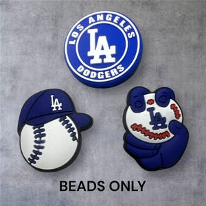 Könnte beinhalten: Drei blau-weiße Perlen im Los Angeles Dodgers-Design. Eine ist ein rundes Emblem mit dem Teamnamen, eine weitere ein Baseball mit einer Kappe und die dritte ein Panda, der einen Baseball hält. Der Text "BEADS ONLY" steht unten.