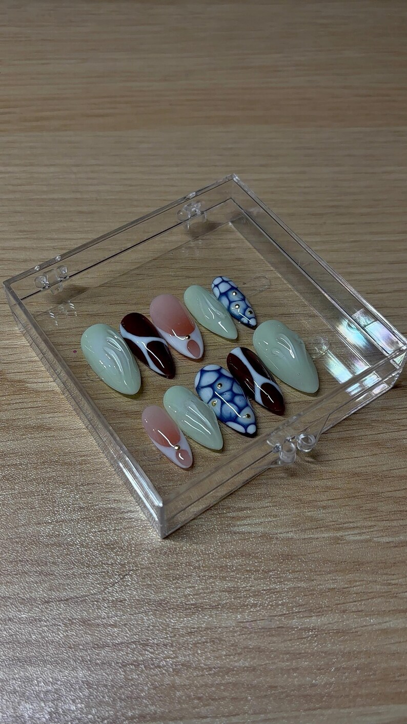 May include: A set of artificial nails in a clear plastic case. The nails are oval-shaped and feature various designs, including solid colors, gradients, and patterned accents. Colors include light green, pink, white, and dark red.