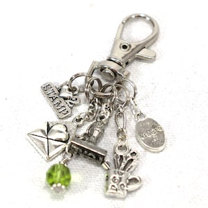 May include: Silver charm keychain with multiple charms, including a green crystal, a stamp, a heart, a letter, a scissors, a cup, and a charm that says "create".