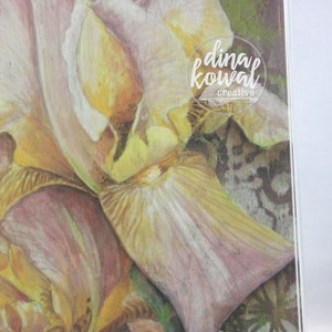 May include: Close-up of a watercolor painting of a pale pink and yellow iris flower. The flower is in focus, while the background is blurred. The artist's signature, "dina kowal creative", is visible in the upper right corner.