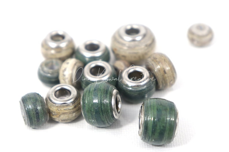Pu&ograve; includere: Primo piano di un gruppo di perline di carta verde e beige con accenti metallici argentati. Le perline sono rotonde e hanno una superficie testurizzata.