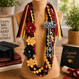 Può includere: Un lei di laurea con nastri neri, rossi e gialli, ornato di fiori di stoffa. Il lei è personalizzato con il nome "LIAM" e l'anno "2026". Sono visibili anche un cappello di laurea e un biglietto con la scritta "Congratulazioni LAUREATO!"
