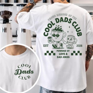 Pode incluir: Camiseta branca com um gráfico verde que diz "COOL DADS CLUB" na frente e atrás. O gráfico nas costas apresenta duas xícaras de café de desenho animado com óculos de sol e o texto "PROUD MEMBER OF THE COOL DADS CLUB EST 2026 POWERED BY LOVE & DAD JOKES".