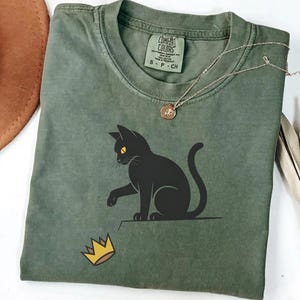 May include: An olive green t-shirt featuring a black cat with yellow eyes reaching for a gold crown. The shirt has a crew neck and short sleeves. A gold necklace with a pendant is also visible.