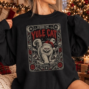 May include: A black crewneck sweatshirt featuring a graphic print of a fluffy cat wearing a Santa hat. The text reads BEWARE THE YULE CAT and GET NEW CLOTHES... OR ELSE! The design includes skulls, stars, and winter foliage in red and white.