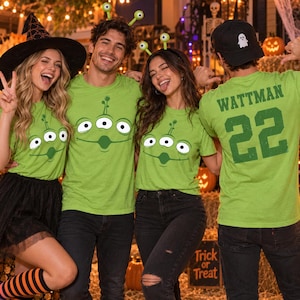 May include: A set of bright lime green t-shirts featuring a printed graphic of a three-eyed alien face. One shirt displays the text WATTMAN and the number 22 on the back in dark green lettering. The shirts are worn with various Halloween accessories.