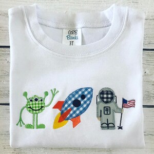 May include: White crew neck t-shirt with an embroidered space-themed design. The design includes a green alien, a blue and white checkered rocket, and an astronaut holding an American flag. The tag reads "ARB Blanks 3T".