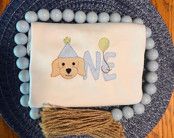 May include: White onesie with a brown dog wearing a party hat and holding a balloon. The onesie says "ONE" in blue letters. The onesie has a brown tassel.