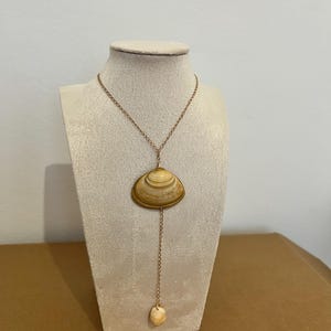 Peut inclure: Un collier de style lasso composé d'une fine chaîne en métal doré. La pièce centrale est un coquillage lisse aux rayures brunes et beiges, avec une perle de coquillage plus petite, arrondie et blanc cassé, suspendue au bout de la chaîne.