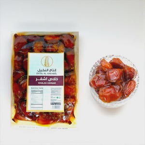 May include: A package of dates labeled "Khalas Ashqar" next to a small glass bowl filled with dates. The dates are a rich brown color and appear to be fresh. The package has a label with Arabic text and nutritional information.