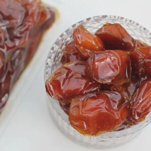 May include: A close-up of a clear glass bowl filled with glossy, brown dates. The dates have a rich, caramel color and a smooth, shiny surface. A portion of dates is also visible in a clear plastic bag.
