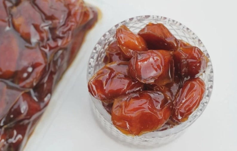 May include: A close-up of a clear glass bowl filled with glossy, brown dates. The dates have a rich, caramel color and a smooth, shiny surface. A portion of dates is also visible in a clear plastic bag.