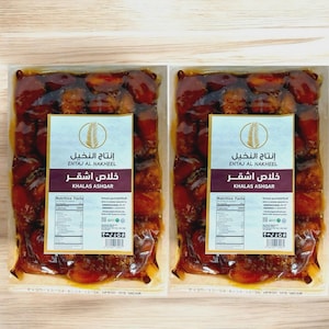 May include: Two packages of Khalas Ashqar dates. Each package is sealed in clear plastic, showcasing the dates' rich brown color. The packaging includes the text "ENTAJ AL NAKHEEL" and "KHALAS ASHQAR" with nutritional information.