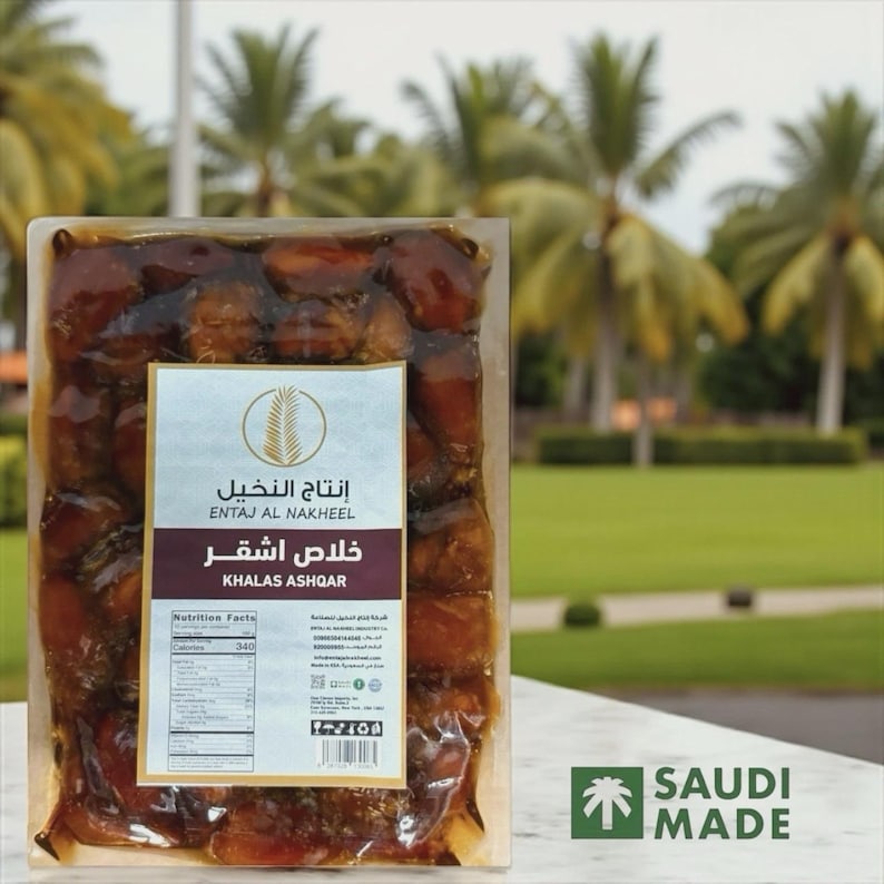 May include: A clear plastic package filled with Khalas Ashqar dates. The label reads "ENTAJ AL NAKHEEL" and "KHALAS ASHQAR". The background features palm trees and a grassy area, with a "SAUDI MADE" logo.