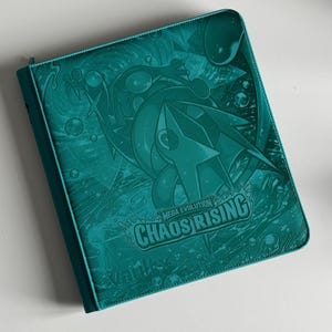 May include: Teal binder with a zip closure. The cover features a detailed embossed design with the text "MEGA EVOLUTION CHAOS RISING". The binder is designed for storing trading cards or other collectibles.
