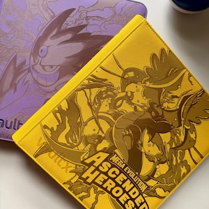 Puede incluir: Dos carpetas para tarjetas, una morada y otra amarilla, con diseños de personajes en relieve. La carpeta morada presenta un personaje con una gran sonrisa. La carpeta amarilla tiene el texto "Mega Evolution Ascended Heroes". Una bola azul y roja está en el fondo.