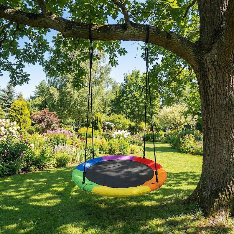 May include: A round tree swing with a black center and a rainbow-colored border of red, orange, yellow, green, and blue. The swing hangs from a thick tree branch with black ropes, set against a backdrop of a lush green lawn and a vibrant garden.