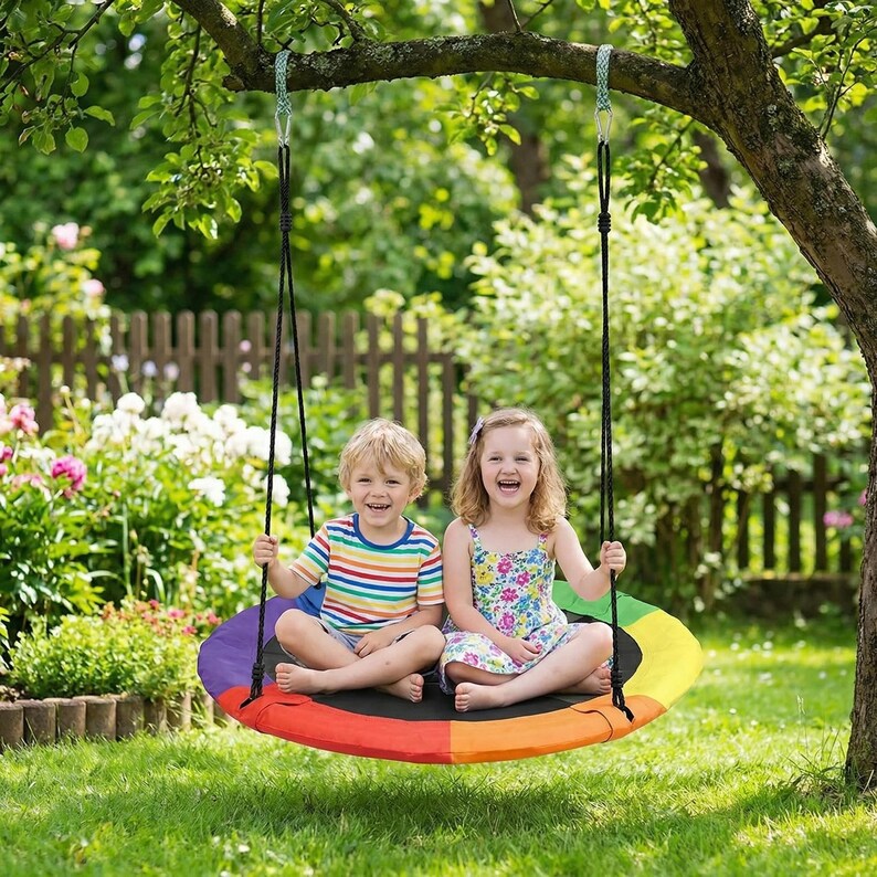 May include: A colorful rainbow-themed tree swing with two children sitting on it. The swing is suspended from a tree branch by black ropes. The children are smiling and laughing in a garden setting with green grass and a wooden fence.