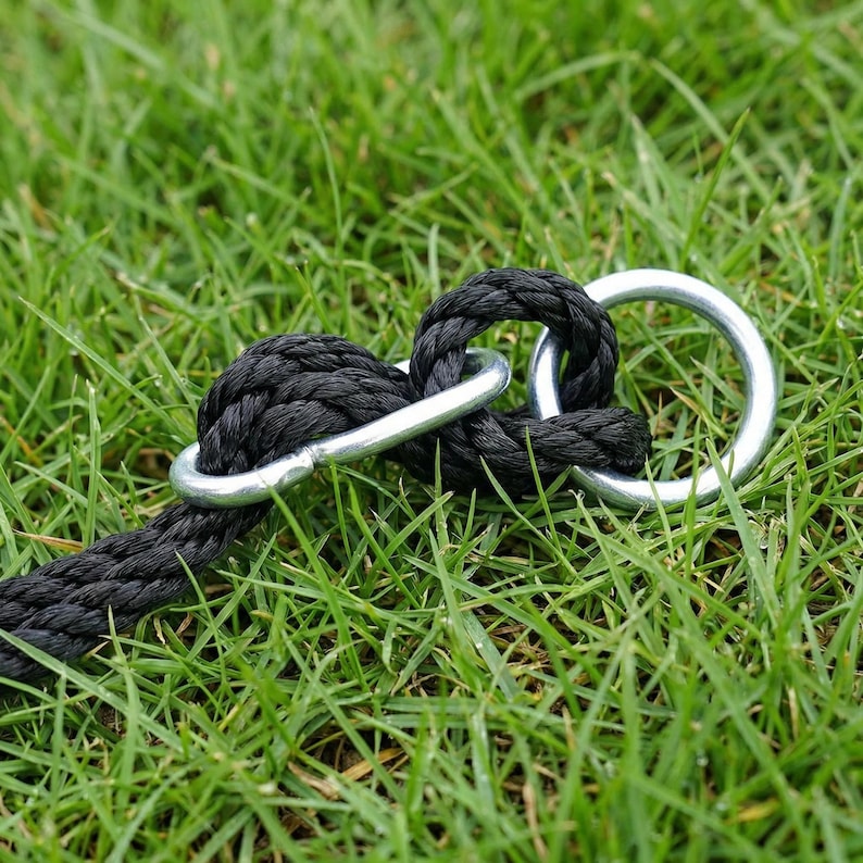 May include: Close-up of a black rope with a loop and two silver metal rings. The rope is thick and textured, and the rings are circular. The items are set against a background of green grass.