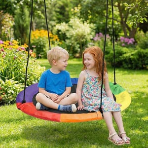 May include: A colorful, round swing with a rainbow design is suspended from black ropes. Two children are seated on the swing, smiling at each other. The swing is set in a grassy yard with a garden in the background.