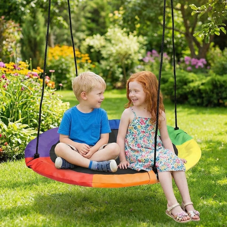 May include: A colorful, round swing with a rainbow design is suspended from black ropes. Two children are seated on the swing, smiling at each other. The swing is set in a grassy yard with a garden in the background.