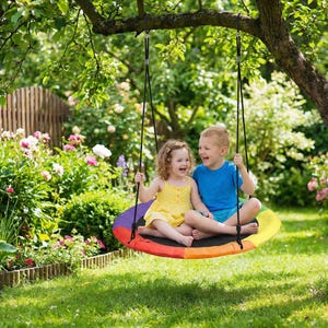 May include: A rainbow-colored round swing hangs from a tree branch. Two children, a girl in a yellow dress and a boy in a blue shirt, sit on the swing, laughing. The swing is suspended by black ropes, and the background features a lush green garden.