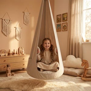 May include: A beige hanging swing chair with a child inside. The swing is suspended from the ceiling and has a soft, rounded shape. The child is smiling and holding onto the swing's handles. The room has a neutral color palette with wooden furniture and macrame wall hangings.