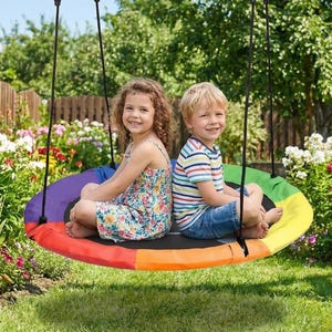 May include: Two children sit on a rainbow-colored tree swing suspended by black ropes. The swing is a large, circular platform with a black center and a vibrant array of colors. The children are smiling, surrounded by a lush green yard.