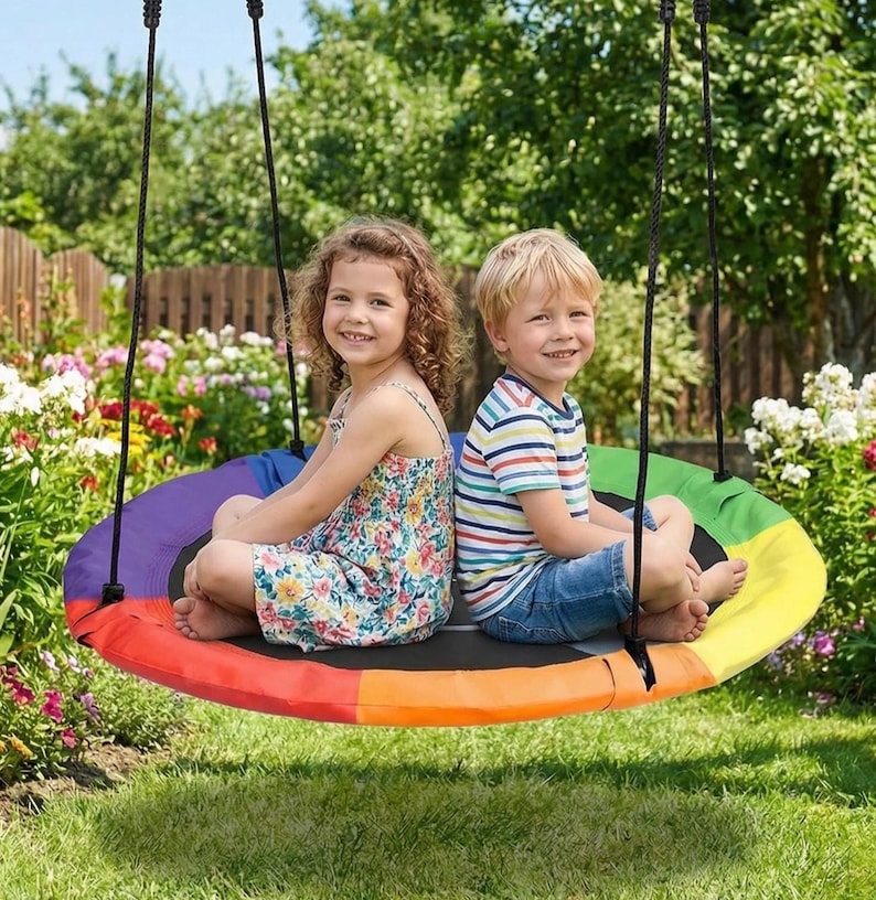 May include: Two children sit on a rainbow-colored tree swing suspended by black ropes. The swing is a large, circular platform with a black center and a vibrant array of colors. The children are smiling, surrounded by a lush green yard.