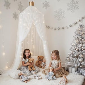 Puede incluir: Dos niñas pequeñas sentadas en una alfombra blanca y esponjosa, rodeadas de juguetes y un árbol de Navidad. Un dosel blanco con una corona dorada encima cuelga sobre ellas, adornado con luces de hadas. La habitación está decorada con copos de nieve y adornos plateados.