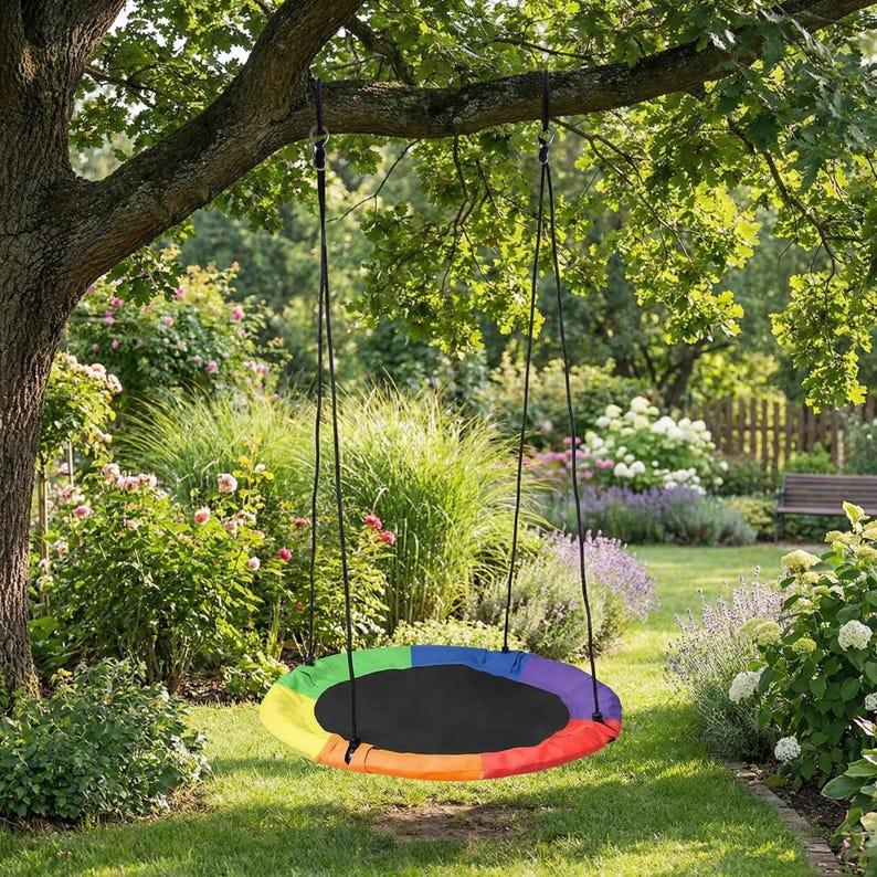 May include: A round rainbow-colored swing hangs from a tree branch in a lush green garden. The swing has a black center and a colorful border of red, orange, yellow, green, blue, and purple. Black ropes suspend the swing.