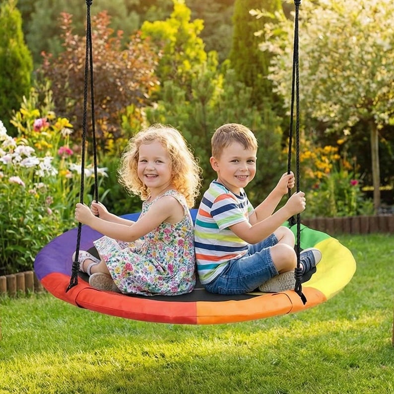 May include: A round rainbow-colored tree swing with black ropes, suspended from a tree. Two children are sitting on the swing, smiling. The swing has a black center and transitions through orange, yellow, green, and purple.