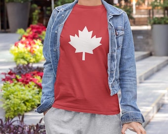 Puede incluir: Una camiseta roja con un diseño de hoja de arce blanca. Se usa con una chaqueta vaquera azul claro y pantalones deportivos grises. La imagen se toma al aire libre, con flores y follaje coloridos en el fondo.
