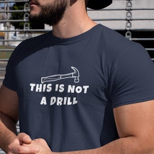 Puede incluir: Camiseta azul marino con el texto "THIS IS NOT A DRILL" y un dibujo de un martillo blanco. La camiseta de manga corta está hecha de un material suave.