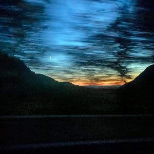 Pode incluir: Uma imagem desfocada de um pôr do sol sobre uma cordilheira, com algumas estrelas visíveis no céu azul.