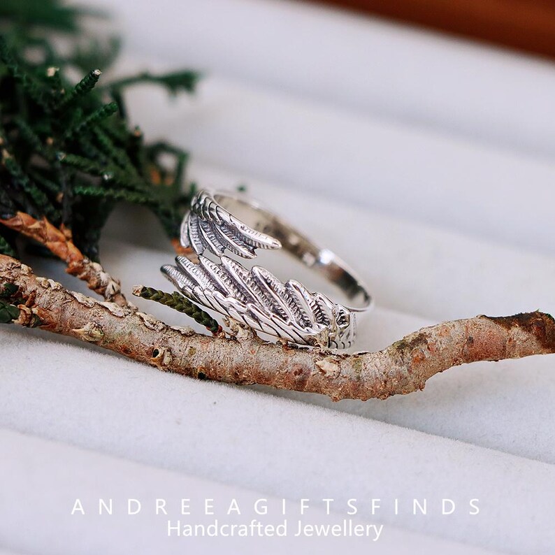 May include: A silver-colored ring with a wing design. The ring is wrapped around a small branch, with green foliage in the background. The ring's wing design is detailed and textured. The text "ANDREEAGIFTSFINDS Handcrafted Jewellery" is at the bottom.